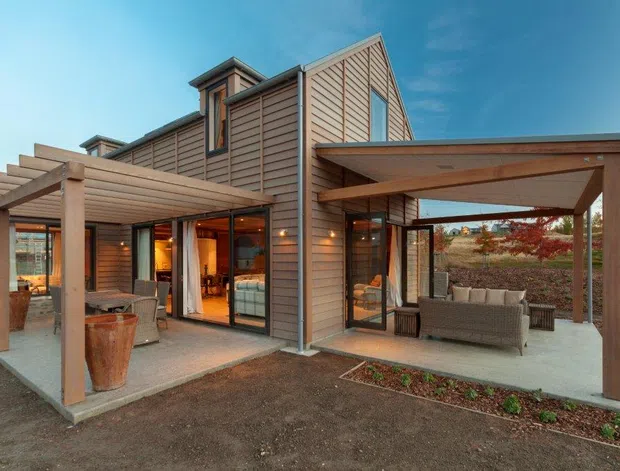 Exterior view of a contemporary multi-level residence with warm horizontal timber cladding, extensive glazing, and two covered outdoor living areas. The structure features exposed timber posts and beams, pergola-style roofs, a steep gabled central volume, and integrated indoor-outdoor entertaining spaces with outdoor seating.