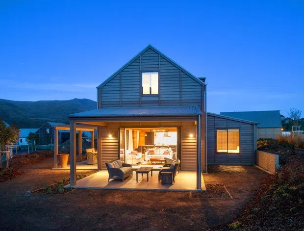 Twilight exterior view of a compact gable-roofed house with dark horizontal weatherboard cladding, a warmly illuminated glazed living area, and a covered outdoor terrace. The home sits on a landscaped site with mountainous terrain visible in the background.