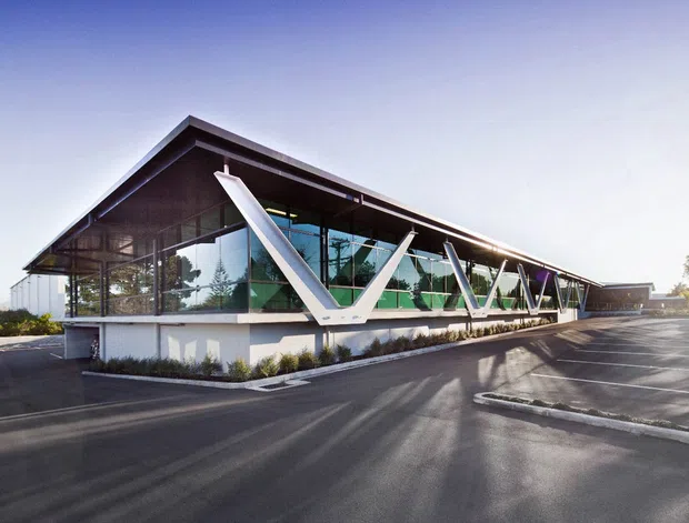 Exterior view of a low-rise contemporary commercial building with a dramatic sloping roof, extensive floor-to-ceiling glazing, and repeated white V-shaped structural supports along the facade. The building sits beside a paved parking area and landscaped edge in bright daylight.