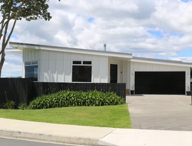 Street-facing view of a contemporary single-storey white-clad house with a low, shallow roofline, large windows, a wide dark double garage, black fencing, concrete driveway and neatly landscaped front lawn.