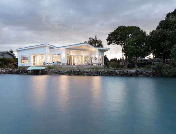 A contemporary single-storey waterfront home viewed from across calm water at dusk. The light-colored house has broad glazing, projecting flat or shallow-pitched roof planes, and a terrace at the water’s edge, surrounded by mature trees and a rocky shoreline.