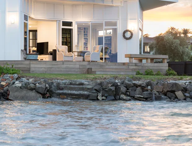 A white, single-storey waterfront house viewed from the water at sunset. The home features a low-pitched asymmetrical roof with broad eaves, white vertical wall panelling, large glazed openings and sliding doors, and a timber deck with outdoor seating. A rocky shoreline and water occupy the foreground, with tropical vegetation around the property.