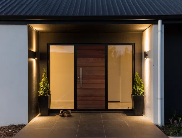 A modern residential entrance viewed from outside, featuring a dark pitched metal roof, white rendered walls, a recessed entry porch with warm integrated lighting, a central timber front door, and tall glazed sidelights. Large dark floor tiles and symmetrical potted topiary frame the doorway.