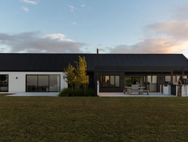 Single-storey modern home viewed from the broad front lawn at dusk. The building has a long dark pitched metal roof, a white rendered wing with large black-framed glazed doors, and a darker covered outdoor entertaining area with built-in barbecue cabinetry. A small landscaped garden and outdoor seating sit along the facade.