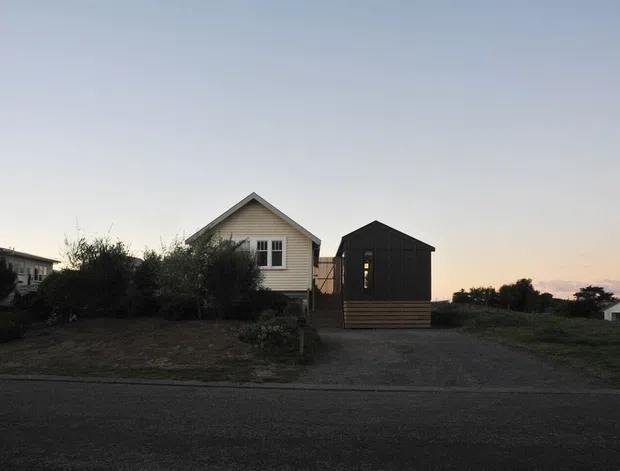 Exterior view of a small coastal-looking residential property at dusk, featuring a light weatherboard cottage with a steep gable roof beside a compact dark-clad gabled structure. The buildings sit on a sparse landscaped lot with a gravel driveway and paved road in the foreground.