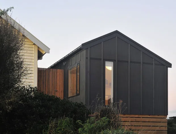 Exterior view of a compact, dark-clad gable-roof building set into a landscaped site. The facade combines vertical dark panels with large screened or glazed openings, while horizontal timber boards form a lower base. A timber fence and the lighter weatherboard side of an adjoining structure are visible at the left.