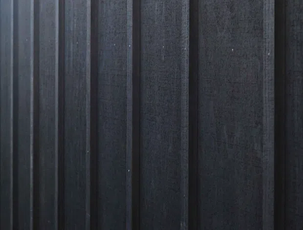 Close-up of a dark, vertically ribbed architectural wall or cladding surface. Narrow raised strips create a regular linear pattern, with a textured charcoal-to-black finish and subtle variations across the panels. No room context or other architectural elements are visible.