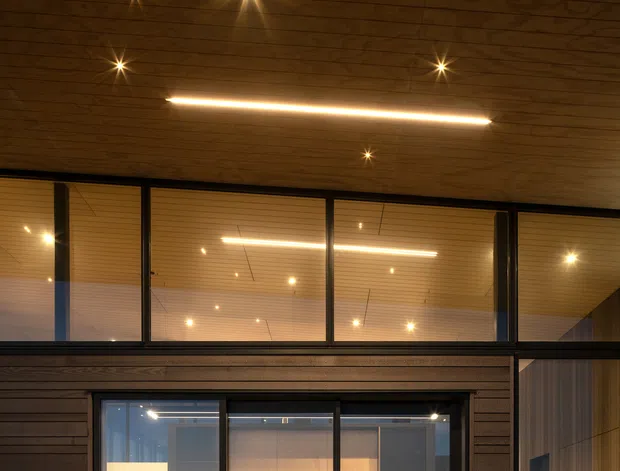 Night-time view of a contemporary timber-clad home with large black-framed glazed openings. A warm wood-lined soffit with recessed spotlights and a linear pendant light projects over the facade. Through the glazing, a compact kitchen or entertaining area is visible with shelving, a benchtop, appliances, and bar stools, while a timber-lined hallway or room is visible at right.