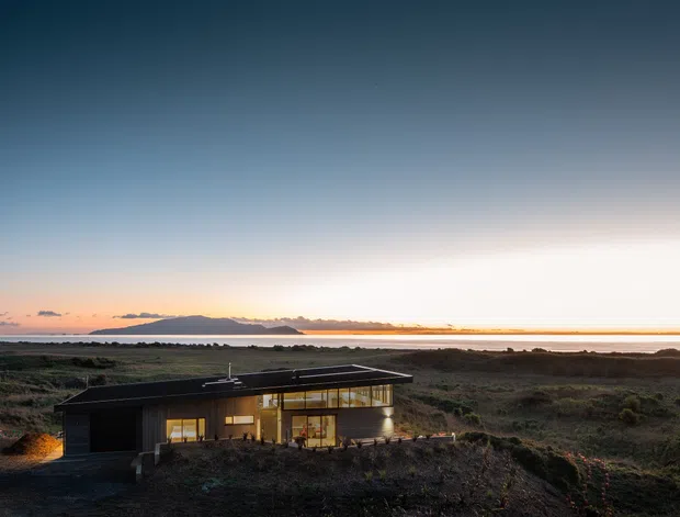 A low, modern single-storey house set within a rugged coastal landscape at dusk. The dark, flat-roofed building has extensive warm-lit glazing and appears integrated into the surrounding scrubby terrain, with a calm ocean horizon and distant landform beyond.