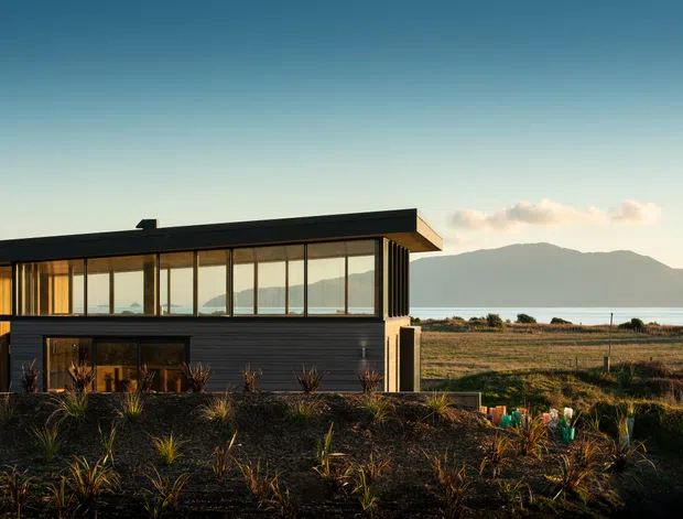 Exterior view of a modern, low-profile two-level home with extensive floor-to-ceiling glazing, dark horizontal cladding, and a flat roof. The building sits in a sparsely landscaped coastal or rural setting, with water and mountainous terrain visible beyond.