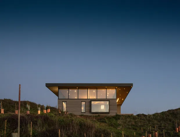 A compact contemporary single-storey dwelling viewed from the exterior at dusk. The building has a strong flat projecting roof with warm recessed lighting, dark horizontal cladding, and a simple rectangular facade with large windows and glazed openings. It sits within a sloping, sparsely planted landscape with protective tree stakes visible in the foreground.