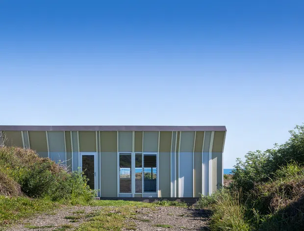A low, single-storey contemporary building set among coastal vegetation. The facade has vertically ribbed or panelled muted green-grey cladding, extensive floor-to-ceiling glazing, dark framed doors and windows, and a shallow flat roof with a dark fascia. A glimpse of blue water is visible beyond the building, while a rough landscaped path and scrub frame the foreground.