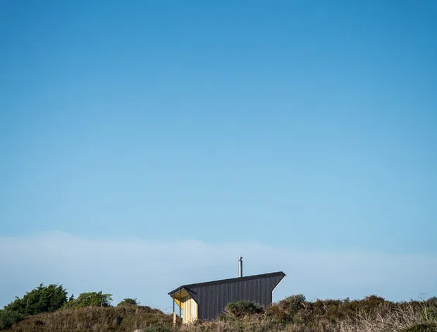 A small, isolated dark-clad cabin or shed is partially visible among coastal scrub and dune vegetation beneath a vast clear blue sky. The simple structure has a sharply pitched asymmetrical roof, a pale timber-colored end wall, and a small vertical chimney or flue. The image is a minimalist exterior landscape photograph with the building occupying a small portion of the frame.