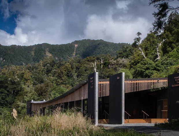 A low, modern building set within dense native forest, with a dramatic mountainous backdrop. The facade combines dark vertical wall panels, tall concrete-like piers, extensive glazing, and warm timber slatted screening along a gently curving roofline. A gravel approach and grasses occupy the foreground, while signage is mounted on the dark entrance wall.