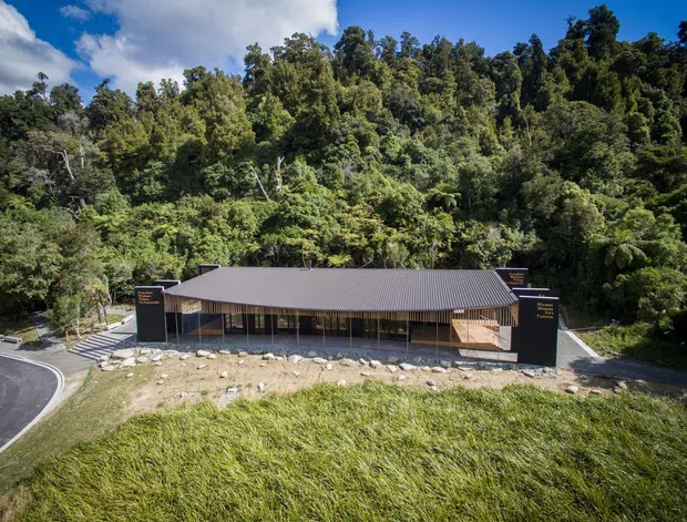 A low, single-storey building viewed from above and at a distance, set against dense native bush. It has a long dark corrugated pitched roof, dark vertical end walls, extensive glazed frontage, and a simple contemporary institutional or commercial form. Curved access road, steps, and landscaped grass are visible in the foreground.