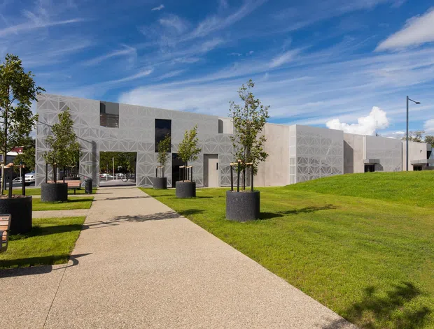 Exterior view of a contemporary low-rise institutional or commercial building set within landscaped grounds. The pale concrete or rendered facade features a repeating geometric relief pattern, deep rectangular openings, and dark window and door elements. A wide pedestrian path leads through lawns with evenly spaced young trees in large cylindrical planters, benches, and simple contemporary lighting. The scene is bright and open under a blue sky.