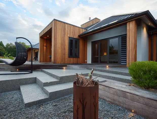Exterior view of a contemporary single-storey residence with warm vertical timber cladding, dark standing-seam metal pitched roofs, deep eaves, large glazed doors, and a stepped textured-concrete terrace surrounded by gravel landscaping. A covered entry recess and landscaped outdoor seating area are visible.