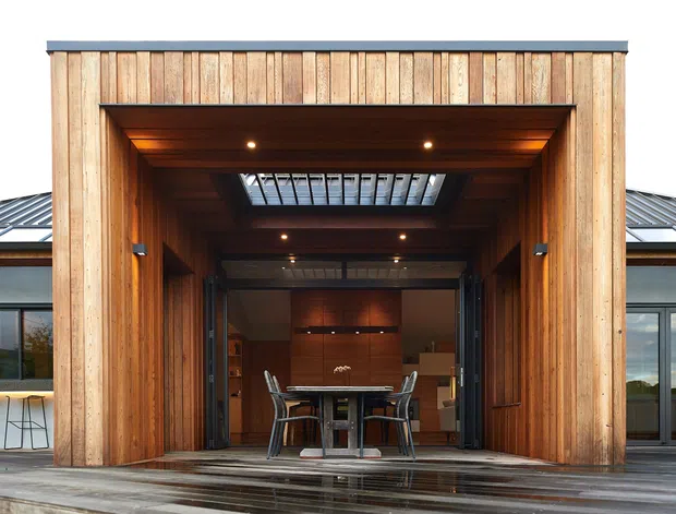 Exterior view of a contemporary single-storey building with a large timber-clad recessed entry or covered outdoor room. The deep timber-lined opening contains a dining table and chairs, with roof glazing or a skylight above, and broad glazed wings extending on both sides. A wet timber deck occupies the foreground.