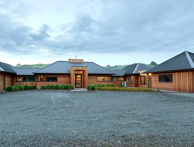 A single-storey residential property viewed from a broad gravel driveway. The low, sprawling home has multiple dark grey metal hipped roofs, extensive warm timber-clad walls, timber-framed windows, a prominent central glazed entry, and an attached double garage. Low planting borders the facade, with open rural-looking surroundings and hills visible in the distance.
