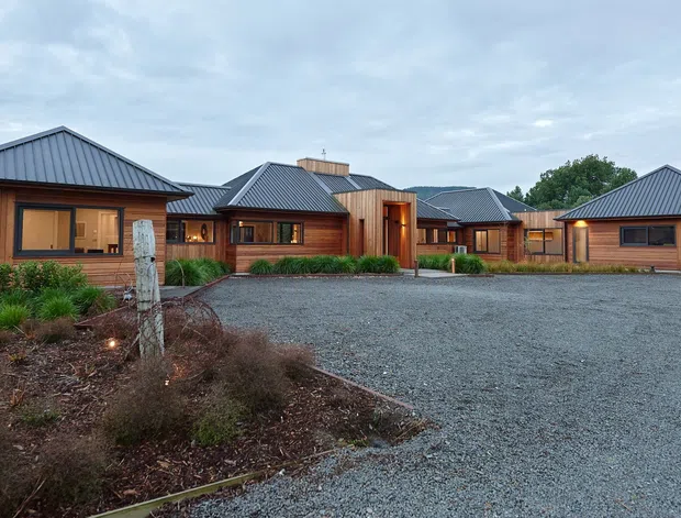 Single-storey residential property viewed from a broad gravel driveway, composed of several connected timber-clad pavilion-like volumes with dark metal hipped roofs. Warm timber window and door frames, landscaped planting beds, and a detached matching garage are visible. Rolling green hills and trees form the distant background under an overcast sky.