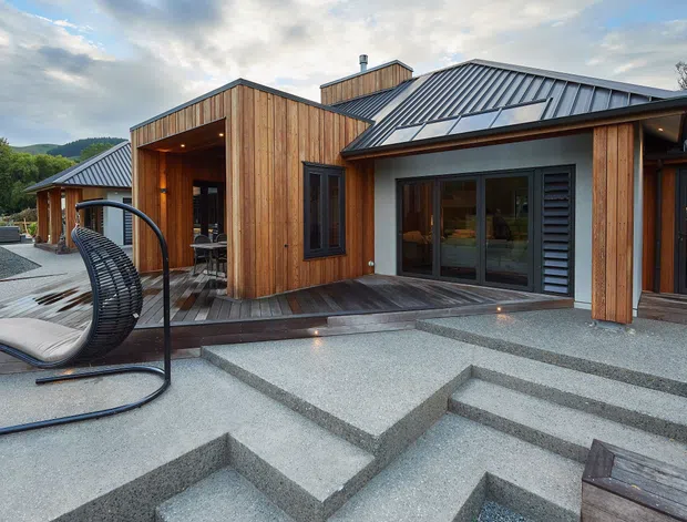 A contemporary single-storey house viewed from an outdoor terrace, with warm vertical timber cladding, dark standing-seam metal pitched roofs, deep eaves, and large glazed doors and windows. Broad geometric concrete steps lead to a dark timber deck and covered entry, with an outdoor hanging chair in the foreground and landscaped rural surroundings beyond.