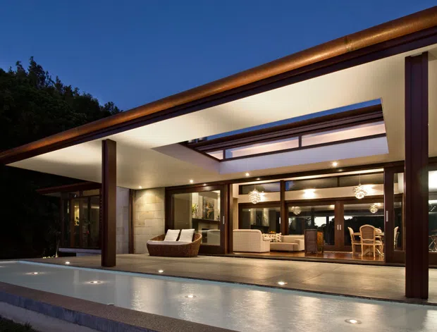 A dusk exterior view of a refined modern single-storey residence, dominated by a broad covered terrace and a shallow reflecting pool in the foreground. Dark timber or stained structural posts frame extensive floor-to-ceiling glazing, while a long linear roof with copper-toned metal edging and a central glazed skylight creates a strong horizontal composition. Warm interior lighting reveals a living and dining area behind the glazing, with recessed ceiling lights and contemporary pendant fixtures.