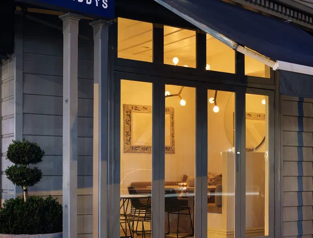 Exterior evening view of a small commercial storefront named Teddy's. The facade features grey horizontal cladding, classical-style pilasters and trim, a dark blue awning, and large glazed double entry doors revealing a warmly lit interior with dining furniture and decorative wall elements.