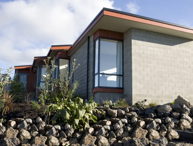 Low-angle exterior view of a contemporary single-storey building with stepped volumes, broad shallow roof eaves, pale concrete masonry block walls, dark window frames, and warm timber-toned soffit and trim accents. The building is set within a landscaped rocky garden featuring dark volcanic-looking stone and planting.