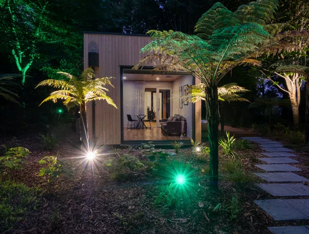 Night-time view of a compact modern pavilion or garden retreat set within dense landscaped vegetation. The dark timber-clad structure has a simple box form, a recessed illuminated entry or living area with large glazing, and a paved stepping-stone path leading through the garden. Landscape lighting highlights ferns and planting around the building.