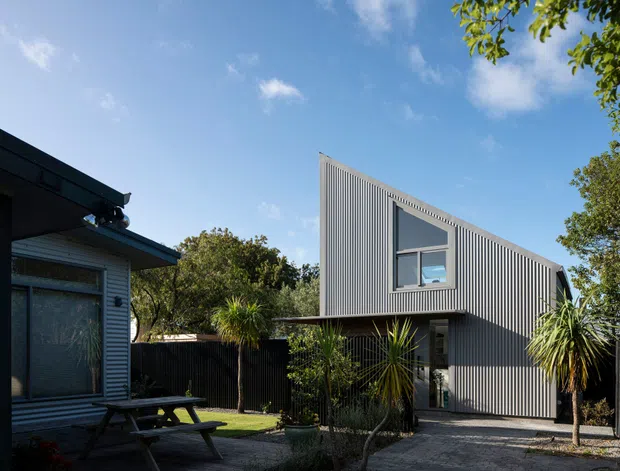 Exterior view of a contemporary two-part residential property. A dark vertically corrugated-clad, steeply sloping gable-like volume sits behind a lower connecting section, with a large upper window and glazed entry. The foreground includes a darker clad structure, paved access, timber picnic table, fencing, and established subtropical planting under a bright blue sky.