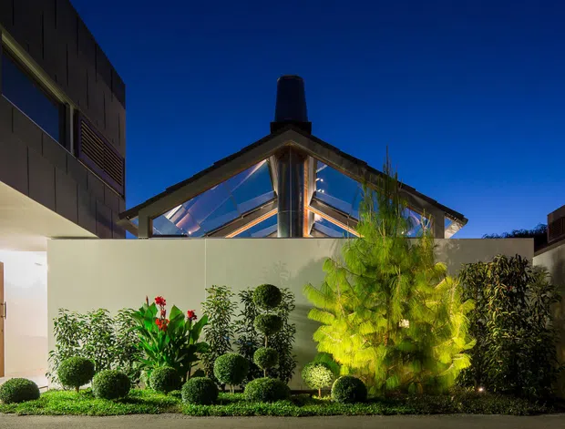 A dusk exterior view of a modern residential property, showing a landscaped front courtyard enclosed by pale rendered walls. The garden features clipped spherical shrubs, layered tropical and ornamental planting, and low groundcover. Behind the courtyard are contemporary multi-level volumes with dark cladding, broad glazing, strong roof planes, and a prominent chimney-like vertical element. Warm architectural lighting contrasts with the deep blue evening sky.