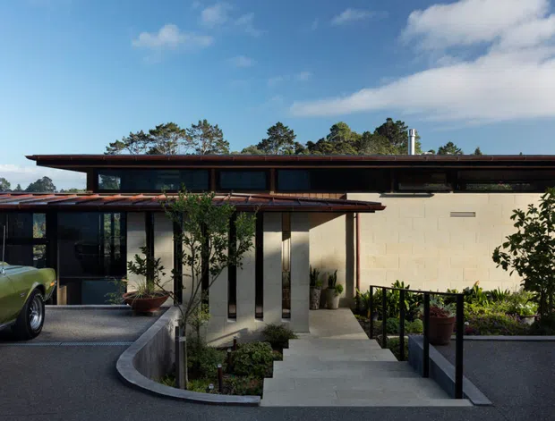 Exterior view of a low, horizontally composed modern house with a broad flat roof, dark metal-framed glazing, pale masonry walls, and a recessed entry approached by concrete steps. The foreground includes a driveway and planted entry garden; a vintage car is present but not part of the architectural classification. The building is set beneath a blue sky with mature trees visible behind it.