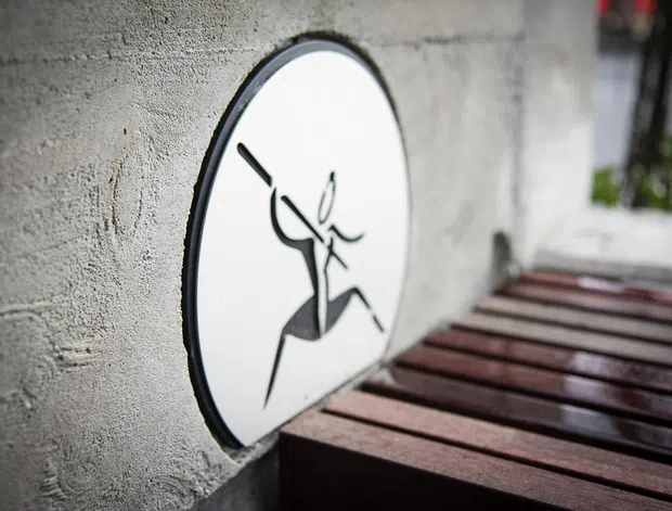 Close-up architectural detail showing a round white sign with a black stylised figure mounted on a rough grey concrete wall. Dark reddish-brown timber slats extend along the lower right, creating a bench or deck-like surface, with a softly blurred exterior background.
