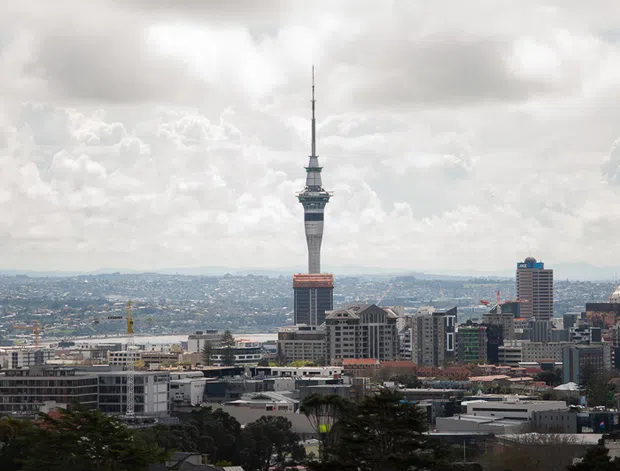 Wide elevated view across a dense urban skyline, dominated by a tall communications tower with a slender spire and observation structure. Mid- and high-rise commercial and residential buildings fill the foreground and middle distance, with low hills and a cloudy sky beyond.