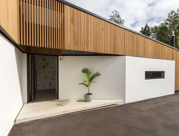 Exterior view of a modern single-storey building with a long, low-pitched roofline, white-painted brick walls, and warm vertical timber cladding forming an upper façade and screened balcony or clerestory area. A recessed glazed entry sits beneath the timber-clad volume, with a narrow horizontal window and paved forecourt. Mature trees are visible in the background.