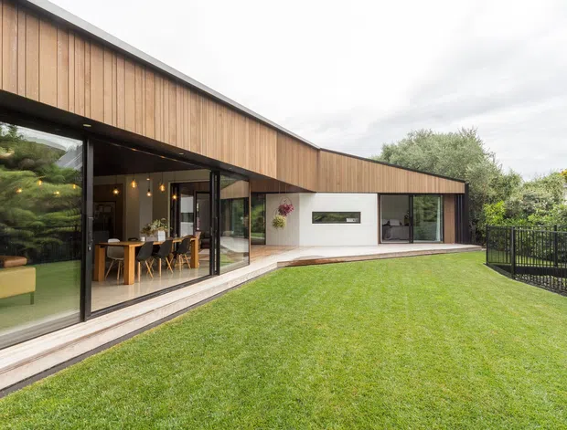 Exterior view of a contemporary single-storey home arranged around a broad lawn and terrace. The building has a long, low profile with warm vertical timber cladding, dark metal window and door frames, extensive floor-to-ceiling glazing, and a projecting roofline. Through the glazing, a dining area and living space are visible. A black metal balustrade borders the right side of the property, with established landscaping around the perimeter.