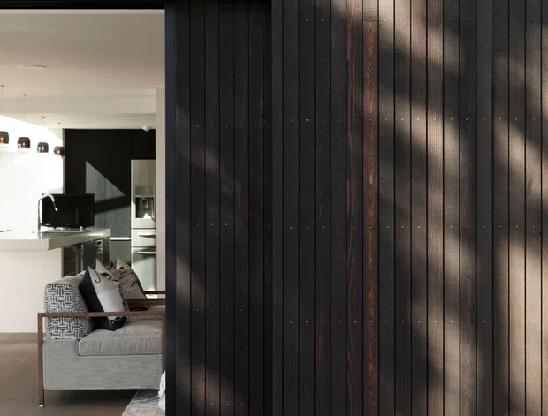 Close architectural view of a dark vertically timber-clad facade with a large black-framed opening. Through the opening, a contemporary living room and kitchen are visible, including a pale sofa, kitchen island, dark cabinetry and pendant lights. Dappled sunlight falls across the timber exterior, creating a warm textural contrast with the dark cladding and metal detailing.
