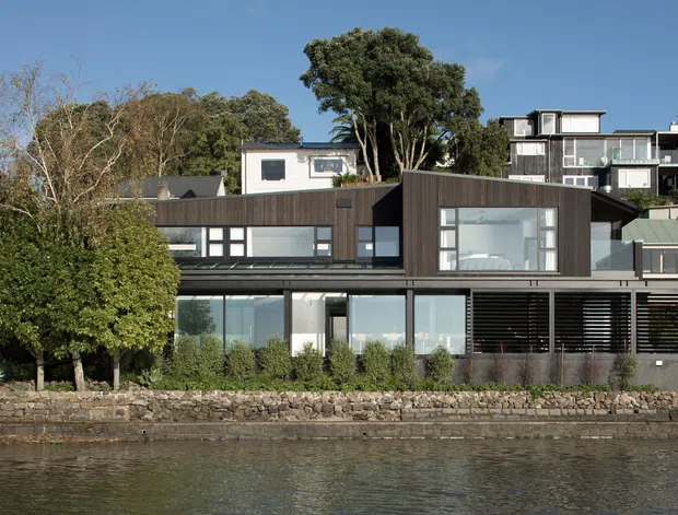 A contemporary multi-level waterfront residence viewed from across the water. The house features dark timber-toned cladding, extensive floor-to-ceiling glazing, horizontal louvres, clean rectilinear volumes, and a stone-edged shoreline with mature vegetation.