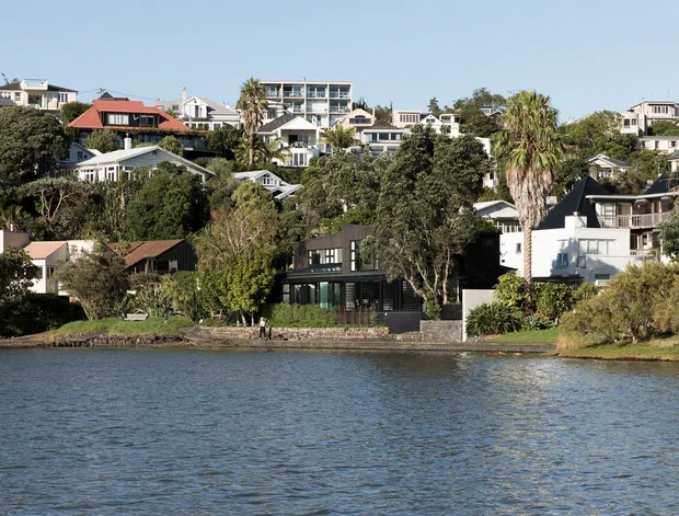 Wide exterior view across a body of water toward a densely landscaped residential hillside, with numerous detached homes and apartment-style buildings. The prominent foreground residence is a dark, contemporary multi-level structure with extensive glazing, set among mature trees and waterfront landscaping. Clear blue sky, palms, and calm water create a bright coastal or lakeside setting.
