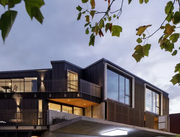A dusk street-level photograph of a contemporary multi-storey mixed-use building. The facade combines dark vertical cladding, extensive glazing, projecting concrete slabs, recessed balconies, black metal balustrades, and warm interior lighting. Commercial or studio spaces occupy the ground floor, with upper-level balconies and windows. Street trees frame the image, and blurred pedestrians appear in the foreground.