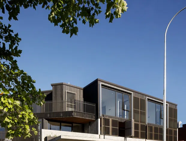 Street-facing contemporary multi-storey building with ground-floor commercial glazing and upper-level residential or office spaces. The facade combines dark vertical slatted cladding, large framed windows, concrete floor slabs, projecting balconies, and metal balustrades. A clear blue sky, mature street trees, and a curved streetlight frame the urban setting.