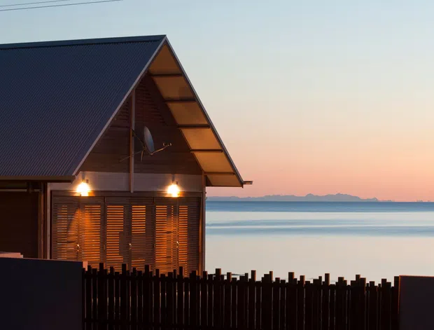 Exterior view of a compact waterside dwelling at dusk, featuring a steep pitched metal gable roof, dark timber walls, deep eaves, exposed timber soffit, louvred timber screening, and warm exterior lighting. The house sits directly beside calm water with a distant shoreline visible beyond, enclosed by a dark vertical timber fence.