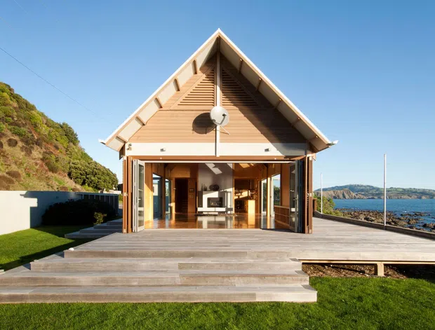Front exterior view of a compact coastal house with a steep front-facing gable roof, timber weatherboard-style cladding, and extensive opened glazing leading onto a broad timber deck and stepped platform. The home sits beside a rocky shoreline with a green sloping hillside and water visible in the background. Warm timber interiors and a fireplace are visible through the open doors.