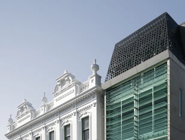 Exterior architectural photograph showing a historic ornate white masonry building adjoining a contemporary dark brick and glass addition. The new structure has a large glazed facade with horizontal sun-shading fins and a perforated dark screen at the upper level, creating a clear heritage-and-modern juxtaposition.