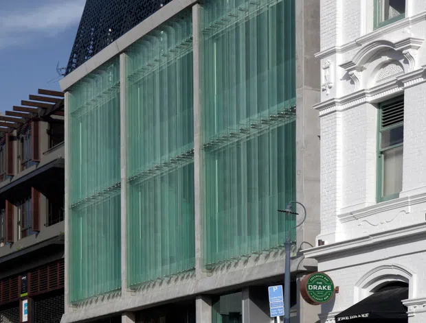 Street-level view of a contemporary multi-storey commercial building inserted beside a highly ornamented white heritage facade. The modern building has a tall glazed curtain wall with vertical green-tinted glass fins, exposed concrete framing, and a perforated dark screen forming the upper facade. Adjacent traditional masonry features elaborate cornices, pilasters, arched openings, and decorative detailing. Urban streetscape with a shopfront and signage at ground level.