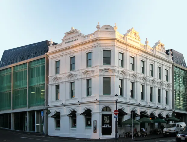 Street-level view of an ornate white heritage commercial building with elaborate classical façade detailing, arched ground-floor openings, decorative cornices, pilasters, and rooftop ornamentation. A contemporary glass-and-metal extension is visible behind and to the left, creating a clear historic-modern contrast. Café seating and umbrellas occupy the footpath at ground level, with surrounding urban streetscape and parked vehicles.
