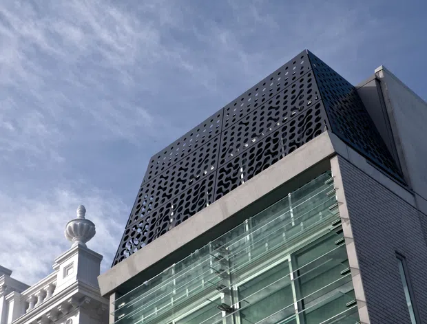 Upward-looking exterior architectural view of a contemporary multi-storey building. The facade combines large transparent glass curtain walls, pale masonry or concrete framing, and a distinctive dark perforated geometric screen at the upper level. A more ornate traditional building is partially visible beside it, creating a contrast between contemporary and historic architecture. Bright daylight and blue sky are present.