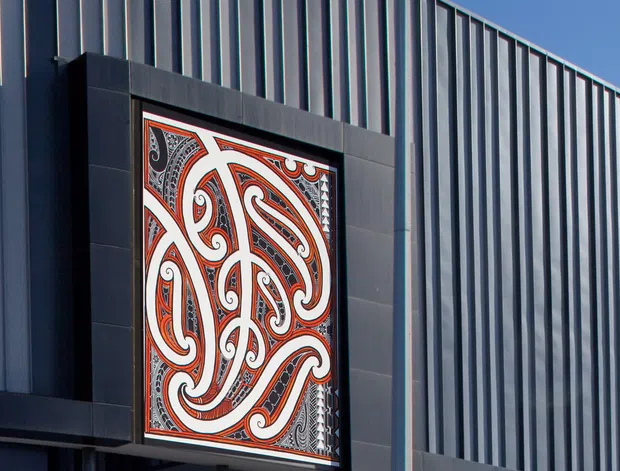 Close-up exterior view of a contemporary commercial building clad in dark charcoal vertical ribbed metal panels. A large framed decorative Māori-inspired artwork with bold black, white, and red curvilinear motifs is integrated into the facade, with partial glazing and structural supports visible below.