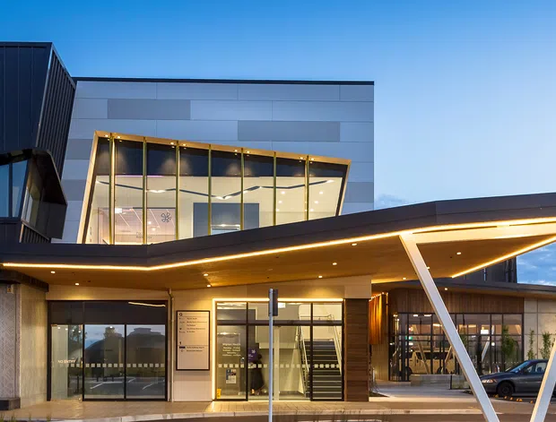Exterior architectural photograph of a contemporary multi-storey commercial building at dusk. The facade combines dark vertical cladding, pale panelled walls, extensive glazed sections, and a dramatic cantilevered entrance canopy supported by angled white structural columns. Warm integrated lighting highlights the entry and canopy, with a glazed upper-level space and urban surroundings visible.