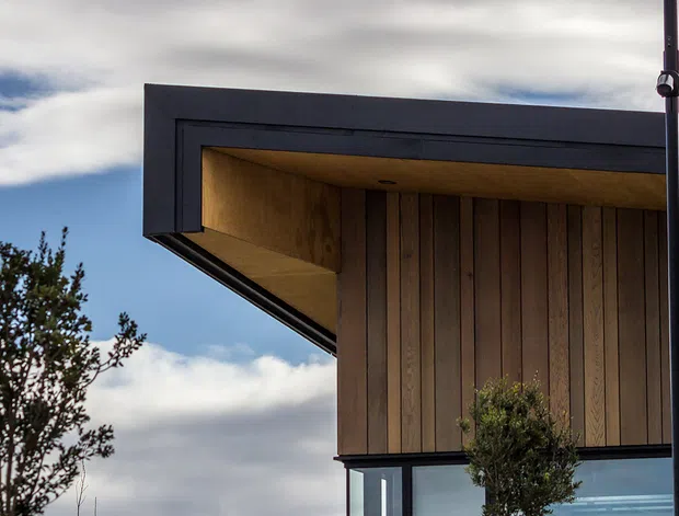 Close-up exterior view of a contemporary building corner featuring a projecting flat roof with dark metal fascia, warm timber soffit and vertical timber cladding, alongside large glazing and a smooth light-coloured wall. Trees and a cloudy sky appear in the background.